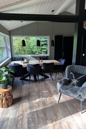 A modern dining area with a wooden table, black chairs, and pendant lights. A large window offers a view of greenery outside. - Bergwild by Interhome (Winterberg)