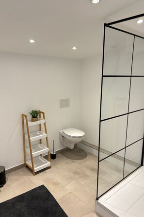 Ein modernes Badezimmer mit Toilette, einem Holzregal mit Pflanze, einer schwarzen Mülltonne und einer Duschkabine aus Glas.
