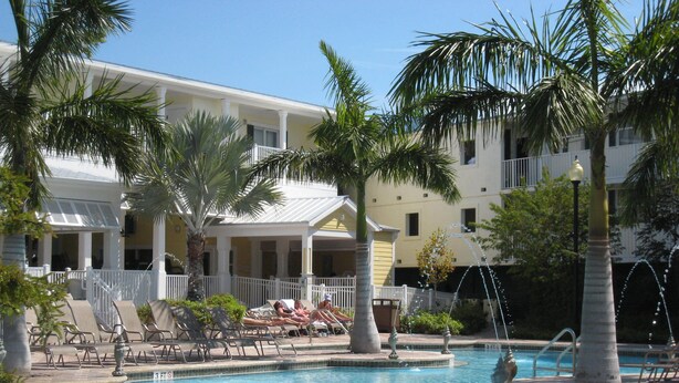 Un área de piscina con sillones de descanso, palmeras y un edificio con balcones.