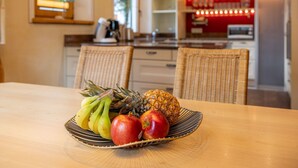 A dining table with a fruit bowl containing bananas, apples, and a pineapple. There are wicker chairs around the table, and a kitchen area with a microwave and shelves in the background. - Villa Emma, Ferienhaus für bis zu 10 Personen, eigener Garten, absolute Privats by Interhome (Kaltenbach)