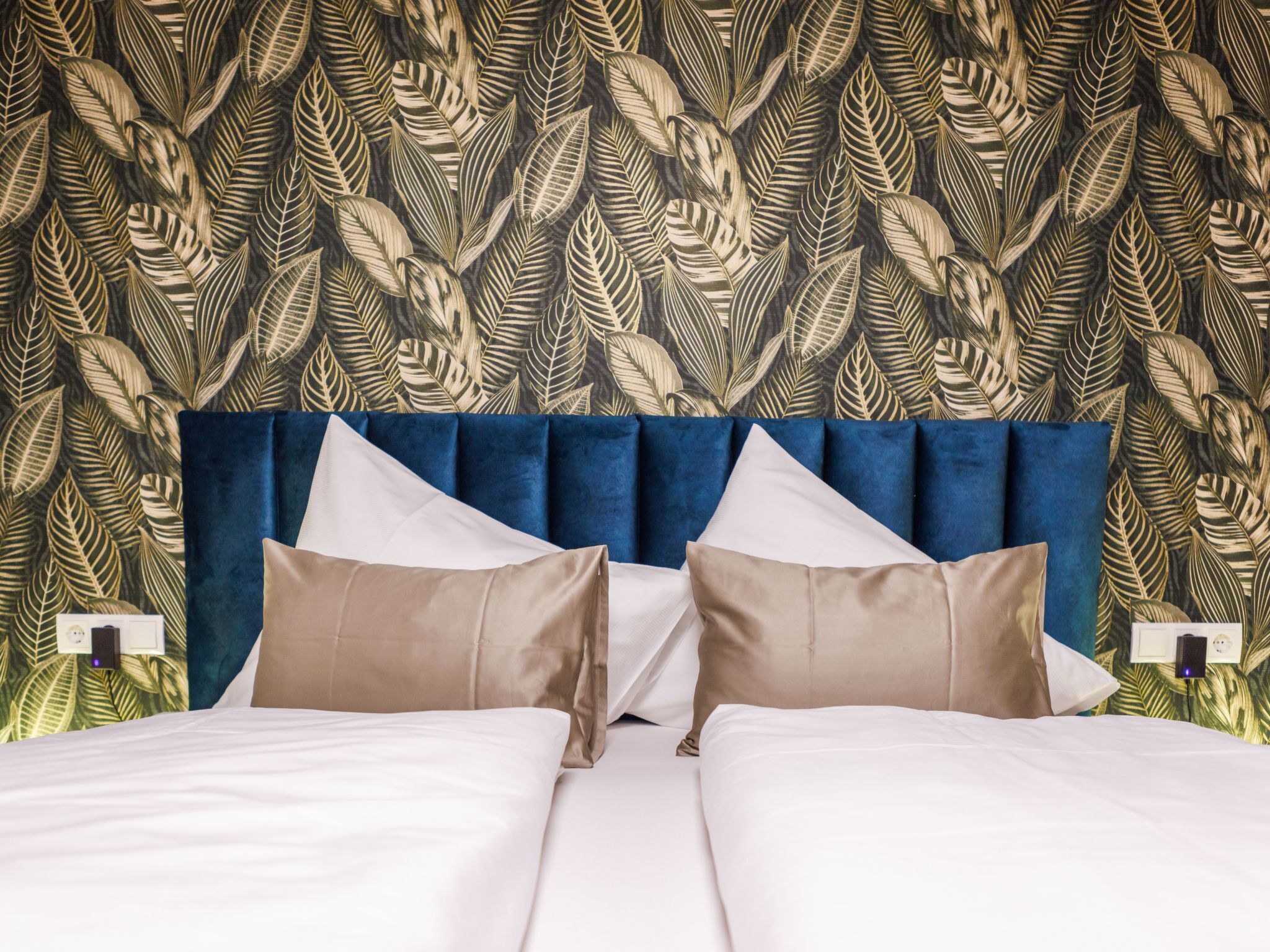 A hotel room with a bed, two pillows, a blue headboard, and a leaf-patterned wallpaper.