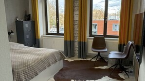Et soveværelse med seng, skrivebord, stol, et oksehudstæppe og udsigt ud
