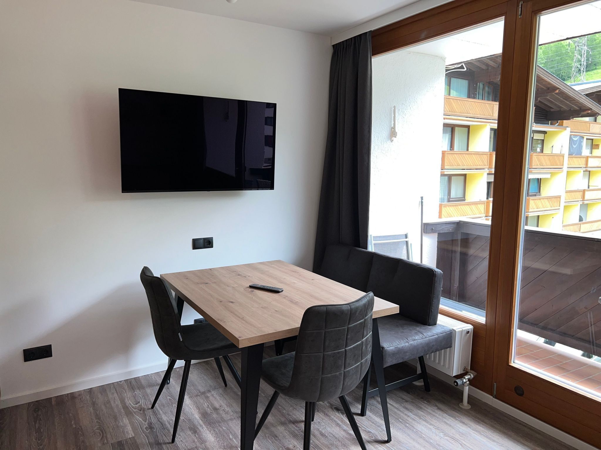 A modern room with a wooden table, two black chairs, a mounted TV, and a large window with a view of a building.
