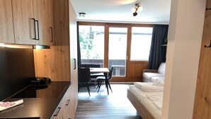 Ett kompakt hotellrum med ett litet kök, en säng, en matplats och ett stort fönster som släpper in naturligt ljus.