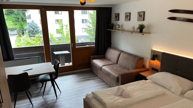Ett hotellrum med en säng, en soffa, ett skrivbord och en stol. Det finns fönster, en hylla med dekorativa föremål och en lampa.