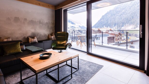 Una sala moderna con un sofá, una mesa de centro de madera y vistas a un paisaje montañoso nevado a través de amplios ventanales.