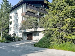 Ein mehrstöckiges Gebäude mit Balkonen, einem zentralen Eingang und umliegender Bepflanzung.