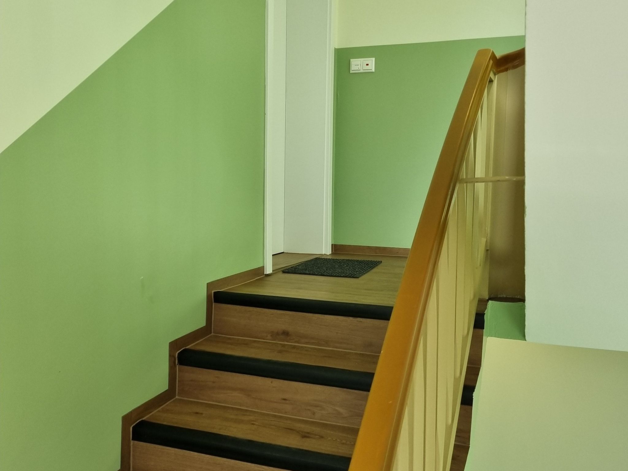 Eine Treppe mit Holzbeschlägen und einem Holzgeländer, die zu einer Zwischenebene mit weißer Wand und Lichtschalter führt.