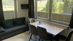 Ein modernes Wohnzimmer mit einer Couch, einem Esstisch mit Stühlen, einem flachbildfernseher an der Wand und großen Fenstern mit Jalousien.