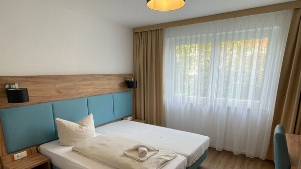 Et moderne hotelværelse med en enkelt seng, træsengegavl, sengeborde, et træskrivebord og et stort vindue med lette gardiner.