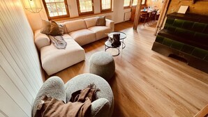 Ein gemütliches Wohnzimmer mit einer Couch, Hocker und einem kleinen Tisch. Es gibt Fenster mit Holzrahmen und einen Kamin.