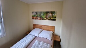 Een slaapkamer met een houten bed, wit beddengoed, een nachtkastje, een wanddecoratie van een bos en een raam met een deur.