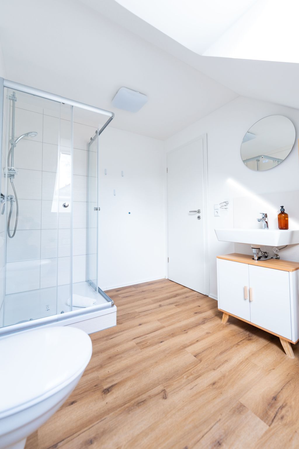 Ein modernes Badezimmer mit einer Duschkabine aus Glas, Holzboden, Waschbecken in Weiß und einem Spiegel an der Wand.