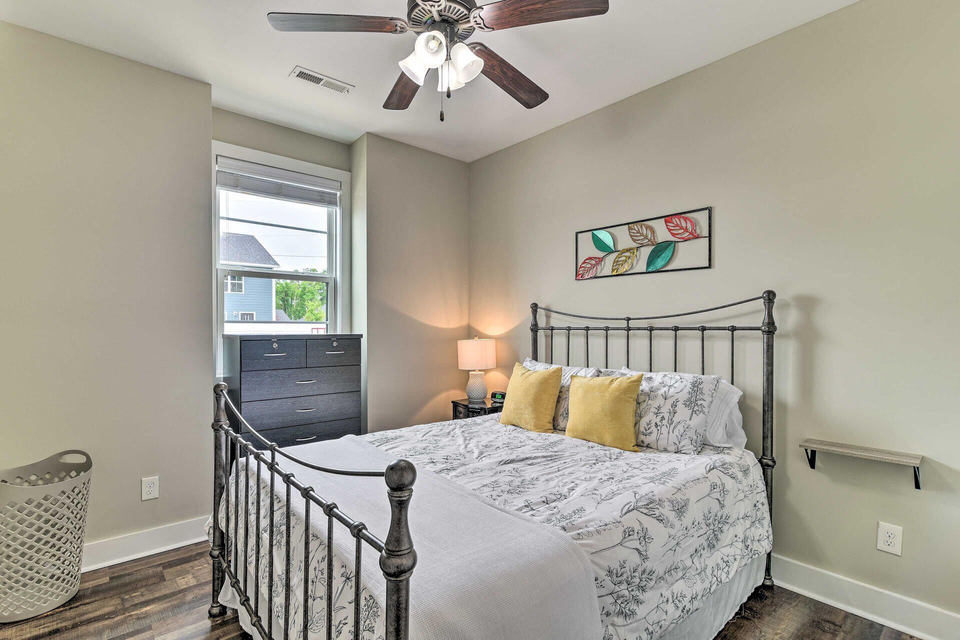 Une chambre à coucher comprenant un lit, une table de chevet, une lampe fixée au mur, un ventilateur de plafond, une fenêtre, une commode et un tableau décoratif représentant des feuilles.