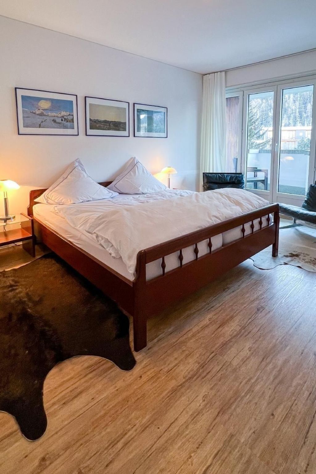 A bedroom with a wooden bed, white bedding, a nightstand with a lamp, a window with curtains, and three framed pictures on the wall.