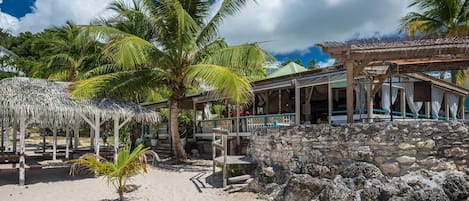 Un resort frente a la playa con muro de piedra, palmeras y una zona de estar techada.