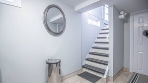 Un pasillo con un bote de basura metálico, un espejo, una escalera y una puerta con letrero de prohibido fumar.