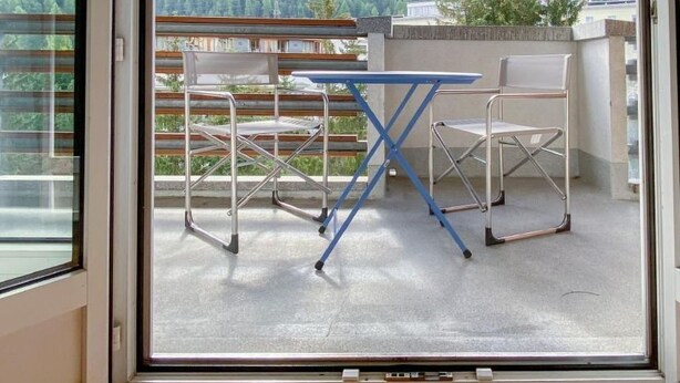 A balcony with a table and chairs, a view of a forested mountain range, and a wooden floor inside. - Derby 37 by Interhome (St. Moritz)