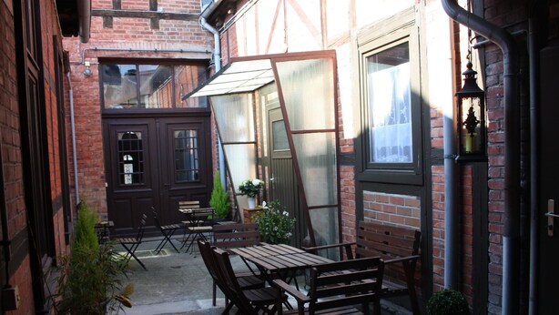 A courtyard with a wooden table and chairs, potted plants, and a brick building with large windows. - in Wernigerode mit Grill und Garten by Interhome (Wernigerode)