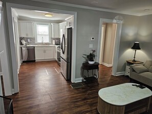 A modern kitchen with white cabinets, a stainless steel refrigerator, and a dishwasher. There is a dining area with a round table and a lamp. - Furnished Atlanta Home Near Airport & Hospitals - Car Available (Atlanta)