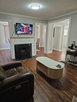 A living room with a fireplace, a television mounted above it, a brown leather sofa, and a wooden coffee table. - Furnished Atlanta Home Near Airport & Hospitals - Car Available (Atlanta)