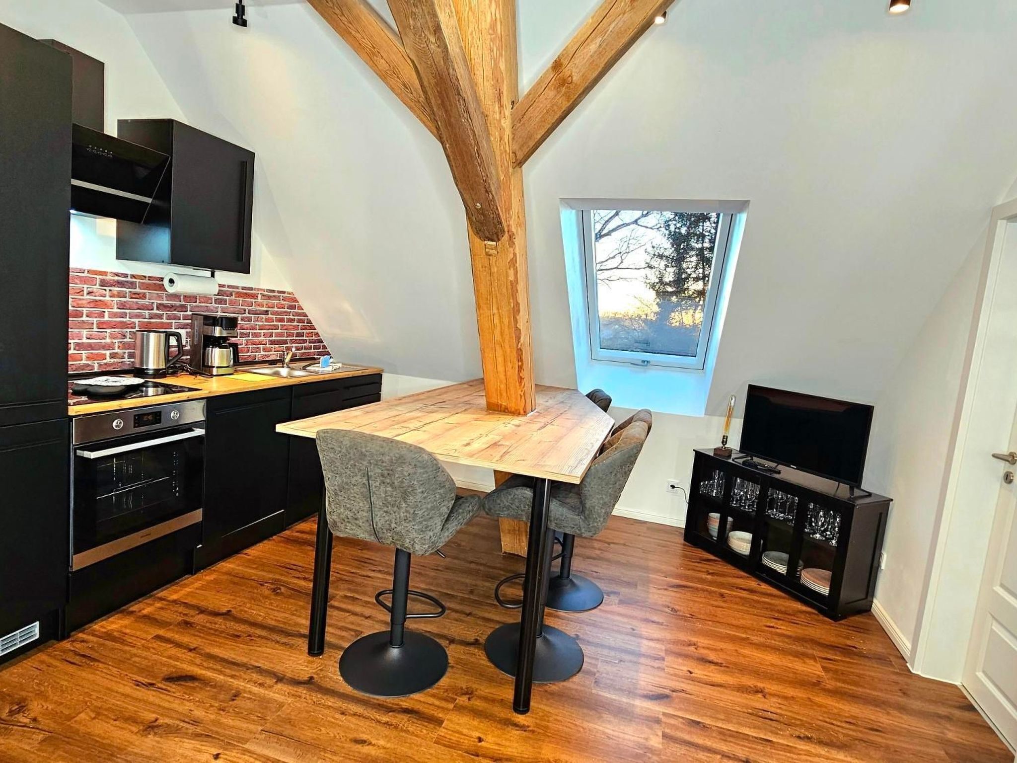 A compact kitchen with a wooden table, two bar stools, a built-in oven, and a TV on a stand.