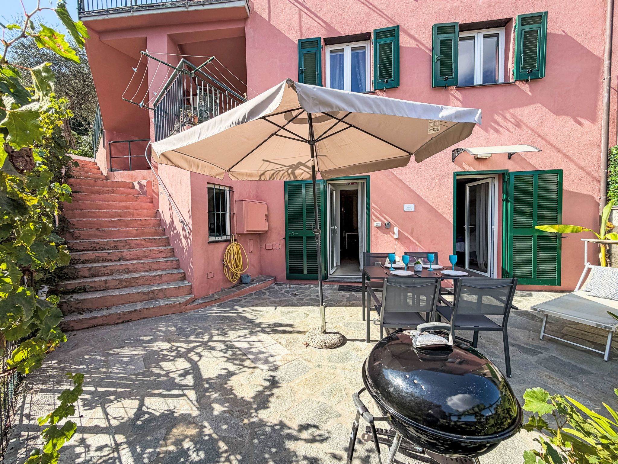 A patio with a grill, outdoor seating, and a staircase leading to a building with green shutters.
