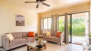 Un salon avec deux canapés, une table basse en verre, un ventilateur de plafond et une porte-fenêtre coulissante donnant sur un jardin.