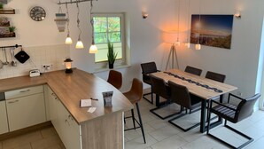 Une cuisine moderne avec un îlot central en bois, une table à manger avec des chaises et des luminaires suspendus. Une fenêtre offre une vue sur la verdure à l’extérieur.