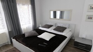 Ein modernes Schlafzimmer mit einem großen Bett, grauen Vorhängen, einem Nachttisch und einer kleinen Nachttischlampe.