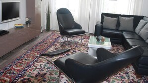 Ein modernes Wohnzimmer mit einem schwarzen Ledersofa, einem schwarzen Ledersessel, einem Glastisch und einem farbenfrohen Teppich mit Muster.