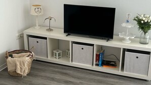 Un salon moderne avec un téléviseur à écran plat posé sur un meuble blanc, des tiroirs de rangement gris, une petite lampe de table, une sculpture d’oiseau décorative, un vase blanc avec des fleurs et un panier en osier.