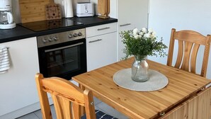 Une cuisine compacte avec des meubles blancs, une cuisinière, une machine à café et un grille-pain. On y trouve une table à manger en bois, des chaises et un vase rempli de fleurs.