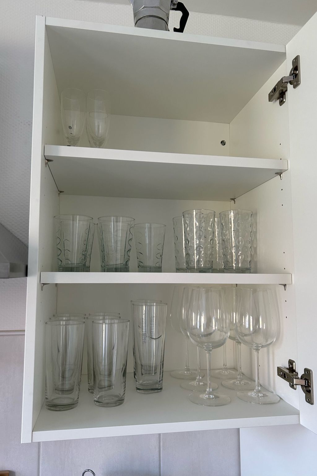 Ein weißes Schrank mit Regalen, auf denen verschiedene Gläser, darunter Tumbler und Weingläser, stehen.