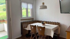 Et spiseområde med et bord dækket af en hvid dug med prikker, omgivet af træstole med hjerteformede udskæringer. En hvid pendellampe hænger over bordet, og et vindue med hvide gardiner giver udsigt til grøn bevoksning udenfor.