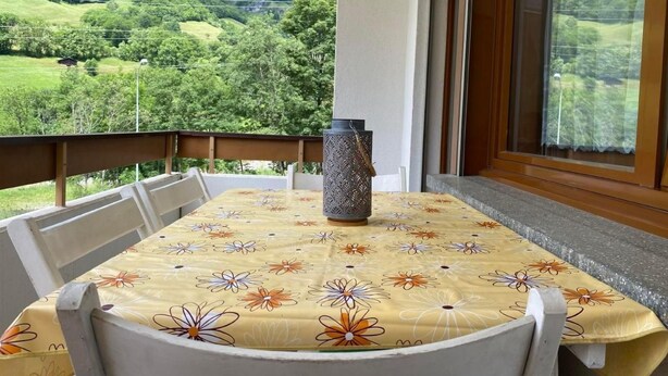 En balkon med et bord dækket af et blomsteret dug, en dekorativ vase og en smuk udsigt over bjergene.