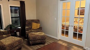 A living room with patterned furniture, a sliding glass door, and a view of a patterned bed. - Cattlemen’s Quarters 3 Bed 2 Bath Apartment (Hebron)