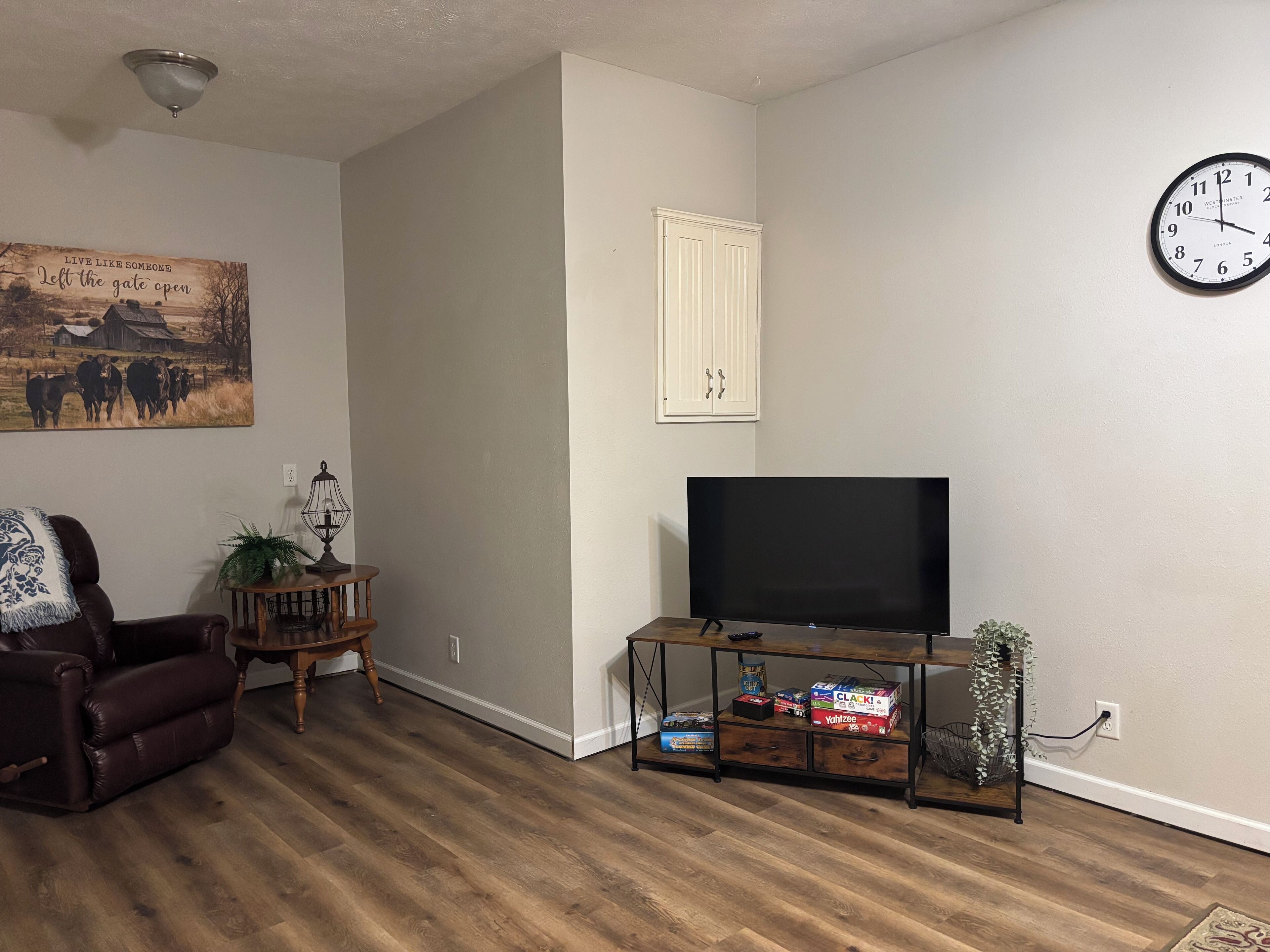 Un salon doté d’un téléviseur à écran plat, d’un canapé en cuir foncé, d’une petite table d’appoint en bois avec une lampe, d’une horloge au mur et d’un tableau représentant des vaches.