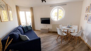 Ett modernt vardagsrum med en mörkblå soffa, ett runt matbord med vita stolar, en platt-TV som är monterad på väggen och ett stort fönster med gardiner.