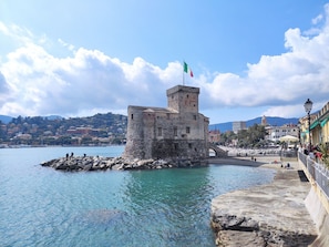 A coastal town with a historic stone tower, clear blue waters, and a paved promenade with buildings and palm trees. - Seestern by Interhome (Rapallo)