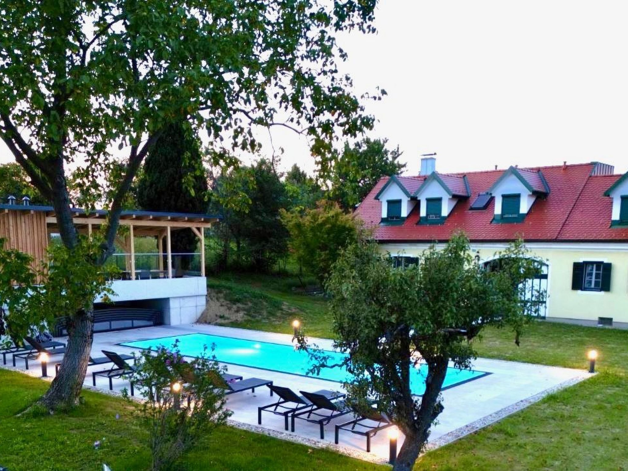 Un área de piscina con sillones de descanso, una terraza de madera y un edificio de techo rojo y múltiples ventanas.