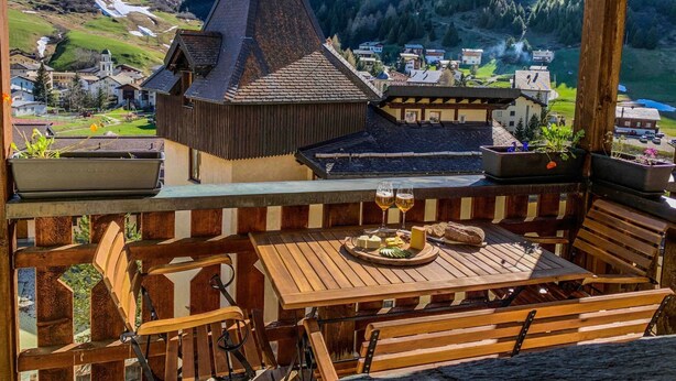 Une terrasse en bois avec une table dressée pour deux personnes, d’où l’on peut admirer un village avec une église et des montagnes en arrière-plan.