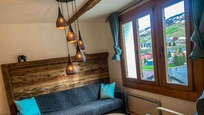 Une pièce chaleureuse avec une tête de lit en bois, un canapé bleu, une table basse en bois et un fauteuil. On y trouve trois lampes suspendues et une fenêtre avec des rideaux.