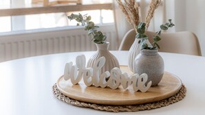 Una mesa redonda con un mantel tejido, un letrero que dice 'welcome' y dos jarrones con plantas secas.