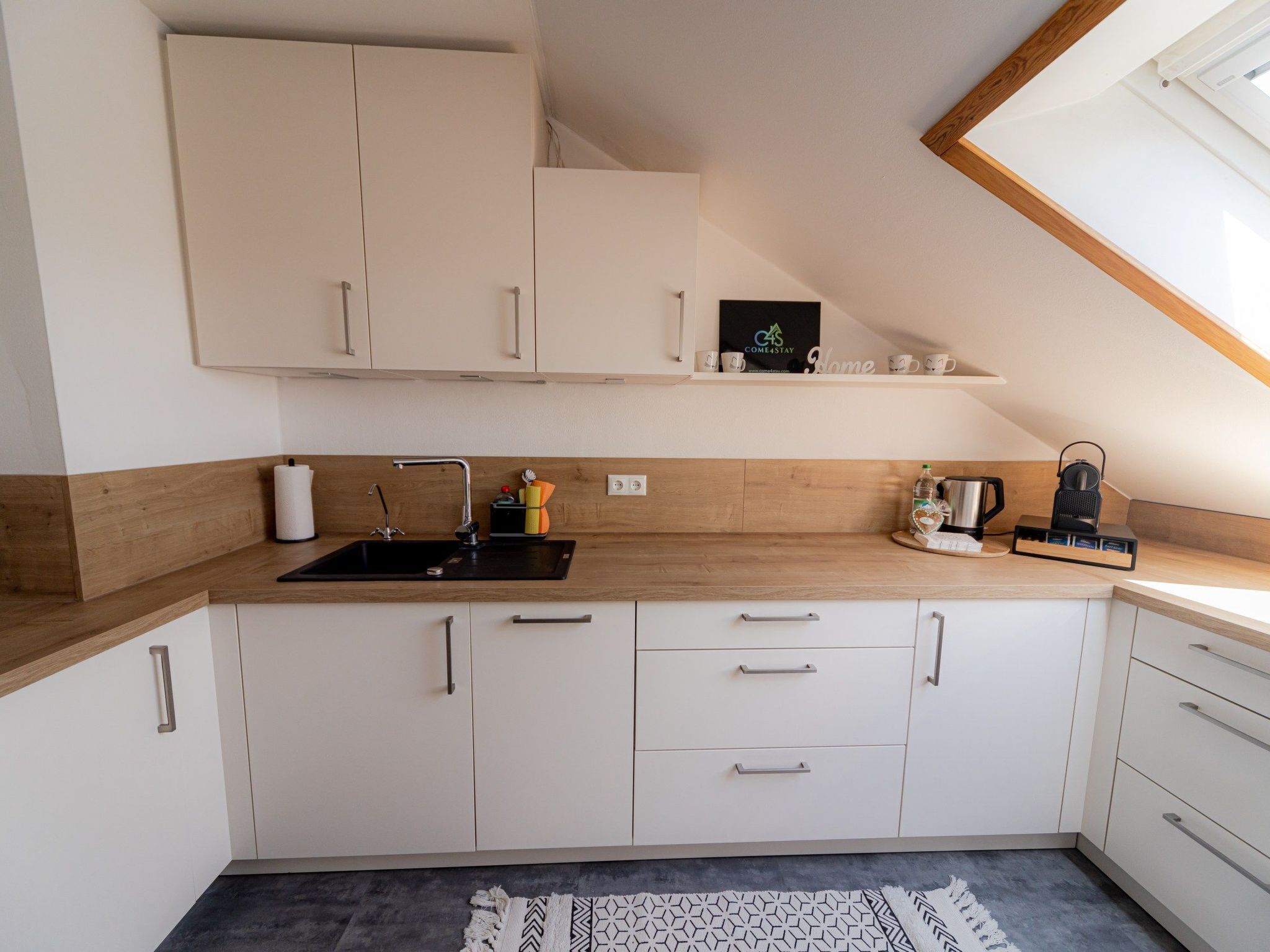A modern kitchen with white cabinets, a black sink, and a wooden countertop. There is a small shelf with a coffee maker and a kettle.