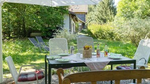 Zona pranzo all'aperto con tavolo apparecchiato per due, circondata da sedie e vista su una casa sullo sfondo.