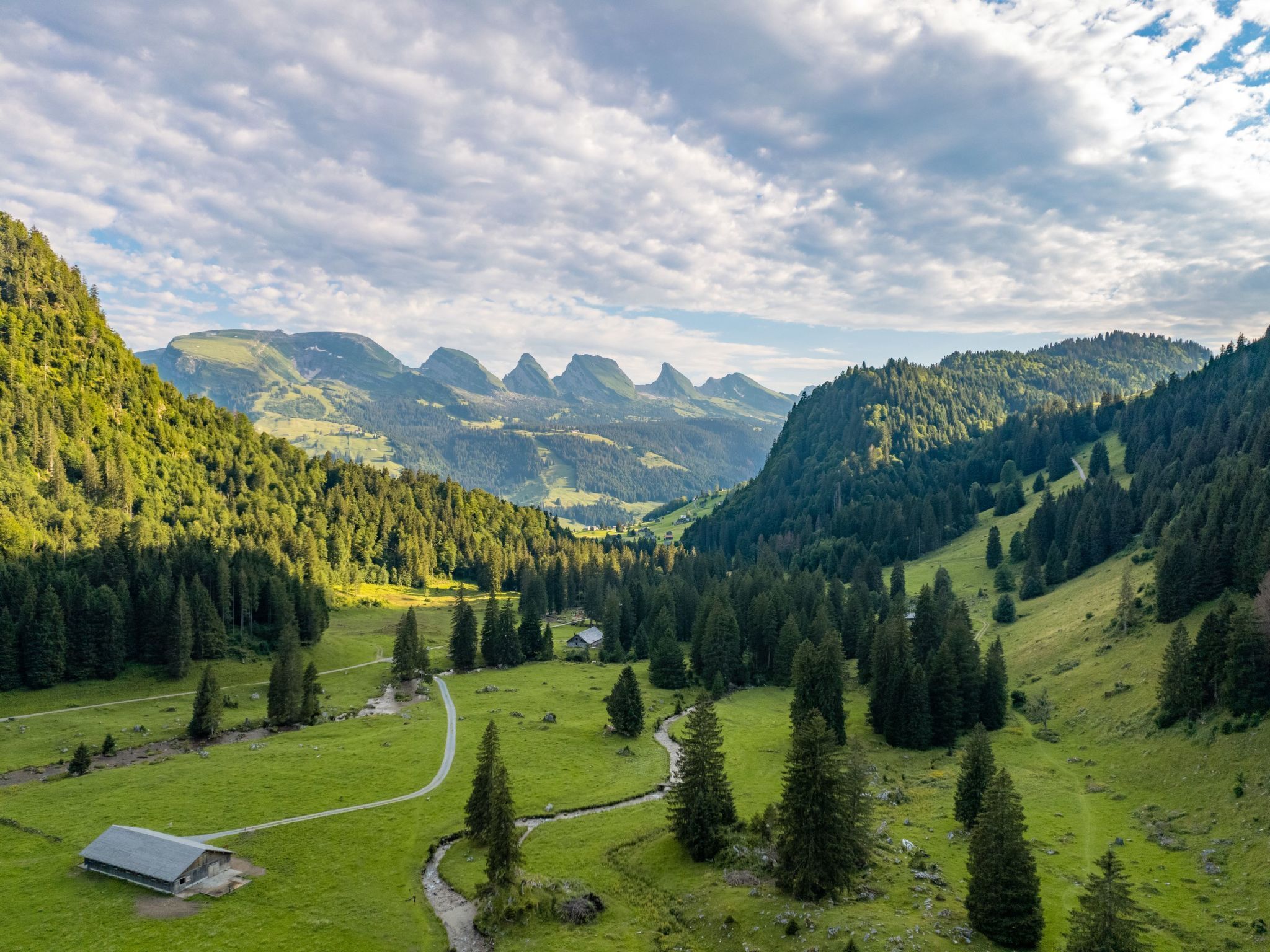 Paisagem rural com um pequeno edifício, um caminho sinuoso e um vale com montanhas ao fundo.