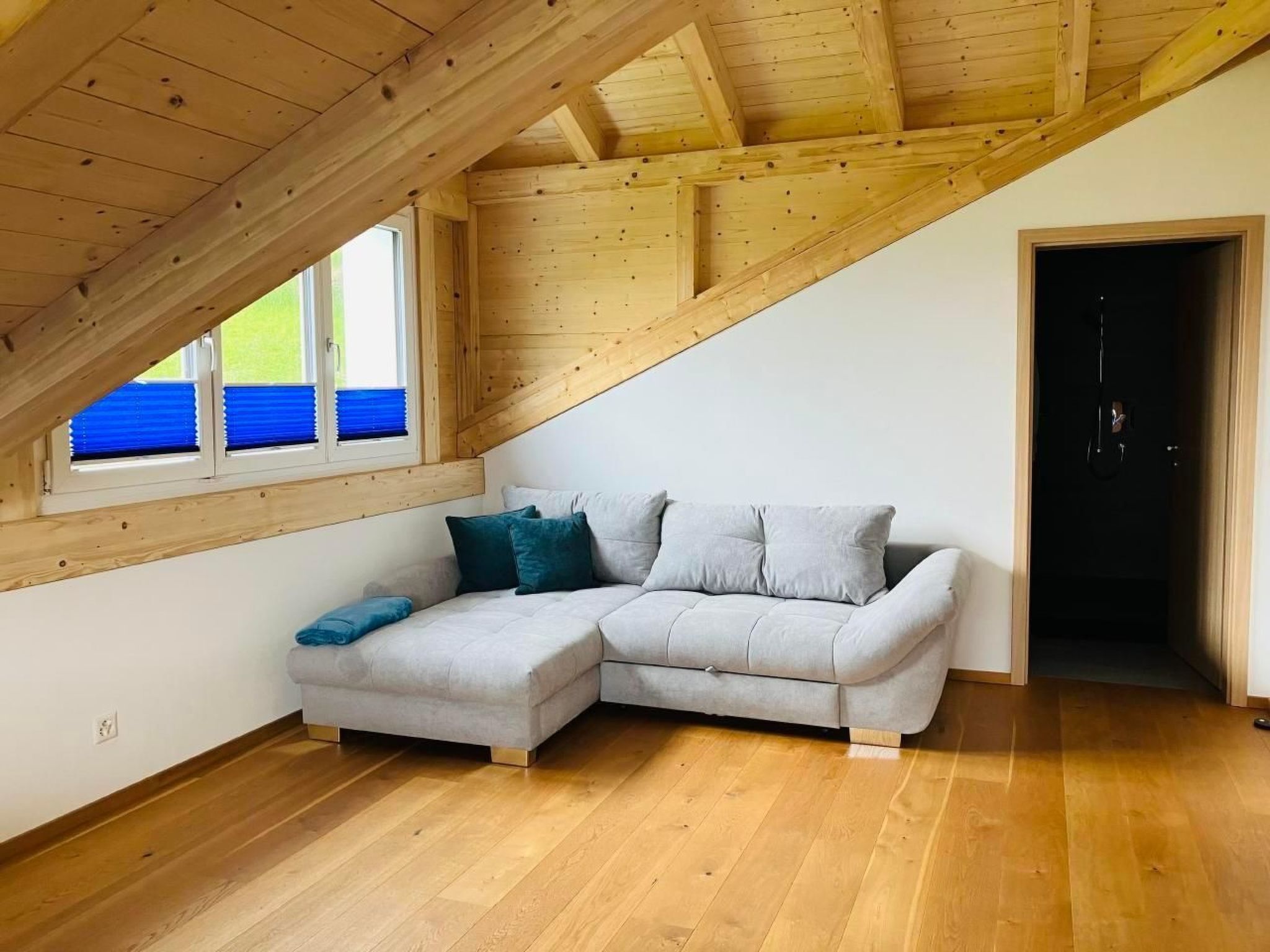 A loft bedroom with a wooden ceiling, a grey sectional sofa, and a window with blue blinds.