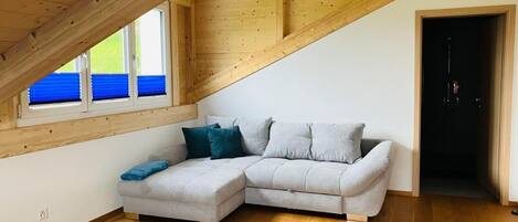 A loft bedroom with a wooden ceiling, a grey sectional sofa, and a window with blue blinds.