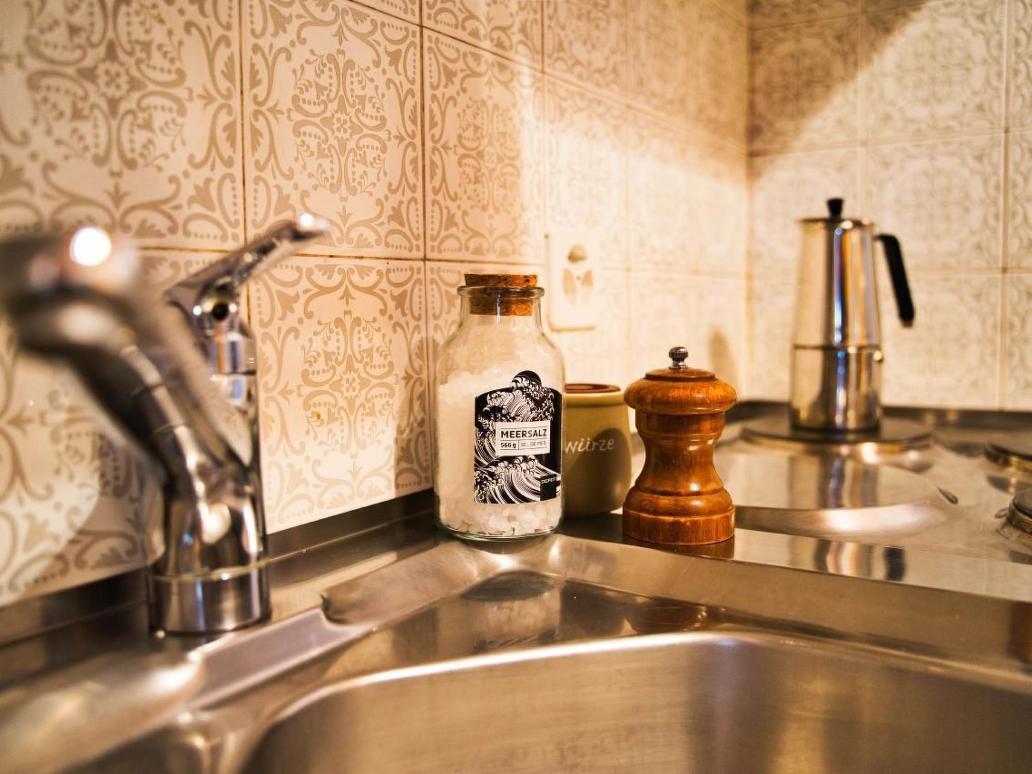 A kitchen with a stainless steel sink, a bottle of Merensalz, a wooden pepper grinder, and a coffee maker.